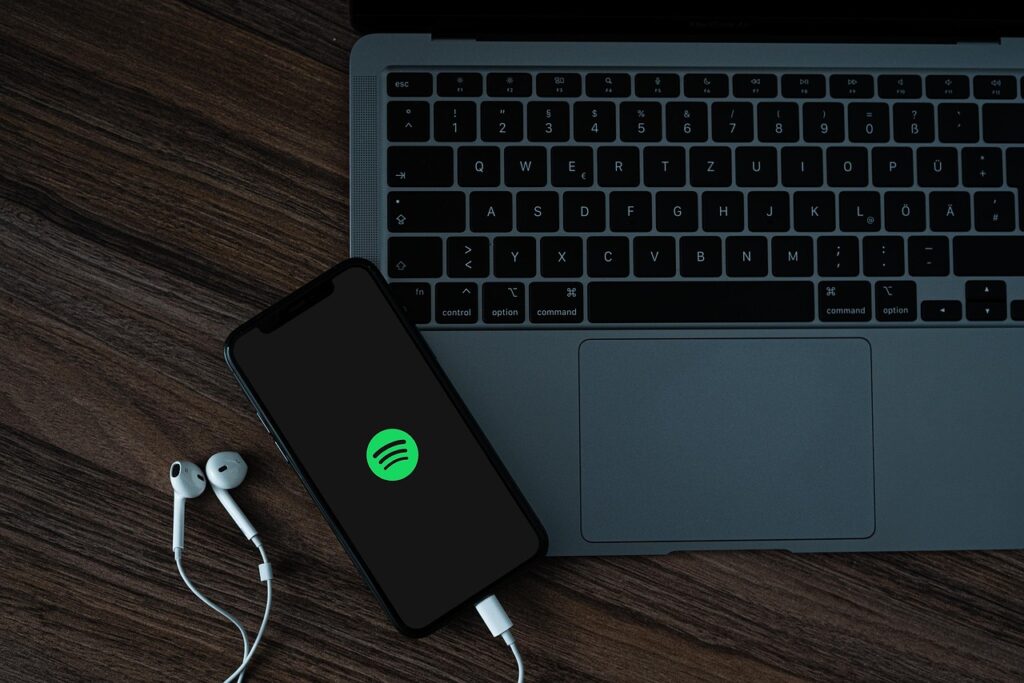 celular com fones de ouvido em uma mesa. na tela mostra a logo do spotify. Na mesa há um laptop.