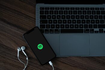 celular com fones de ouvido em uma mesa. na tela mostra a logo do spotify. Na mesa há um laptop.