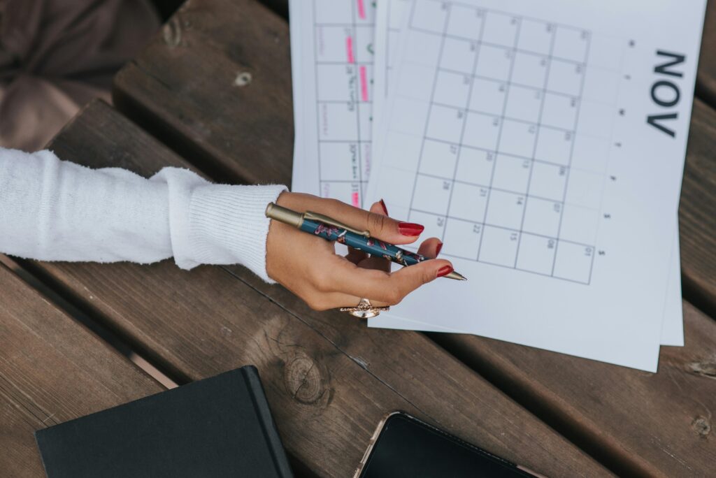 folha a4 em formato paisagem com o calendário de novembro de 2024. Uma mulher está com a caneta na mão e com o braço apóiado em uma mesa rústica de madeira. Há também um telefone celular e uma agenda ao lado. O rosto dela não aparece na imagem.